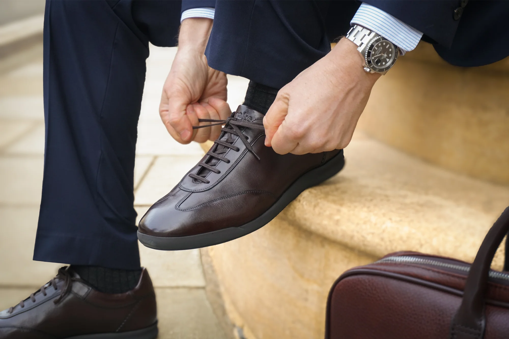 A man in a navy blue suit sitting on a step wearing his Loake Mayfair Smart Trainers in Dark Brown Leather.