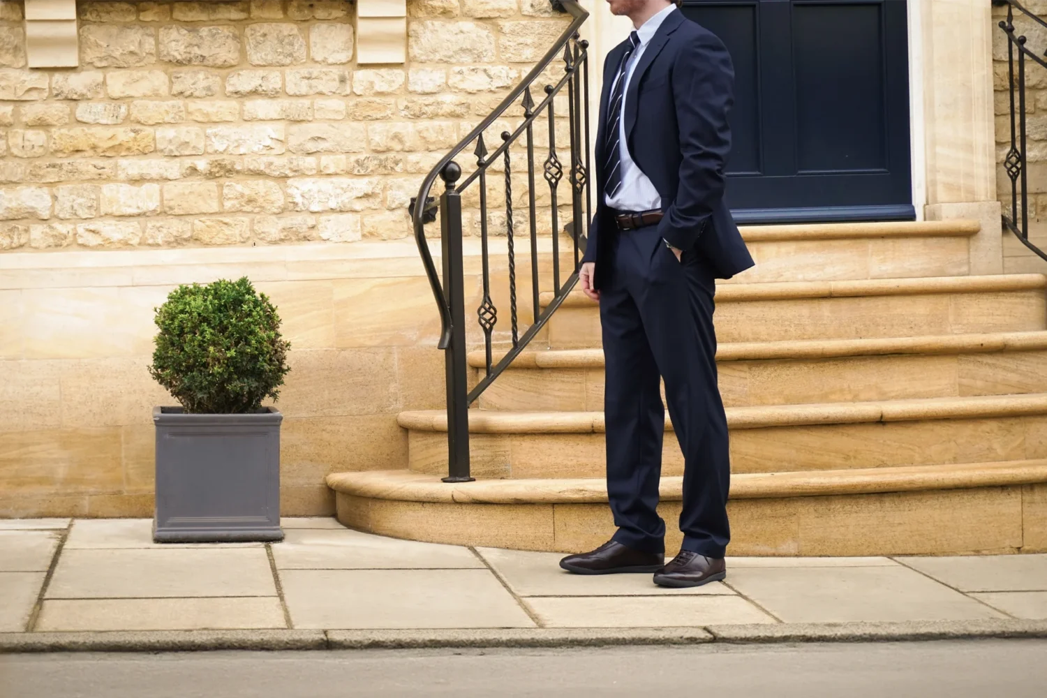 A man in a navy blue suit standing in the street wearing his Loake Mayfair Smart Trainers in Dark Brown Leather.