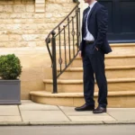 A man in a navy blue suit standing in the street wearing his Loake Mayfair Smart Trainers in Dark Brown Leather.