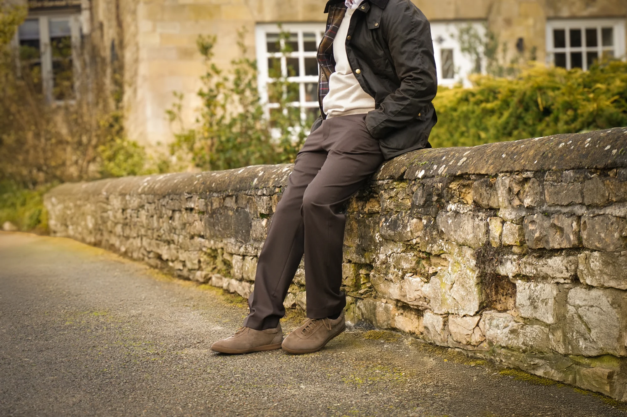 A man in smart brown trousers and brown coat standing in the street wearing his Loake Mayfair Smart Trainers in Flint Suede.
